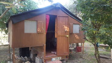 A small wooden cabin stands elevated on stilts, built atop a trailer. Surrounded by trees, its door is open, revealing a red curtain inside.