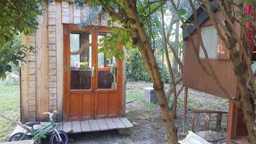 A small wooden cabin with glass doors stands in a garden, surrounded by trees. Nearby, a child's green and purple bicycle rests on the grass.