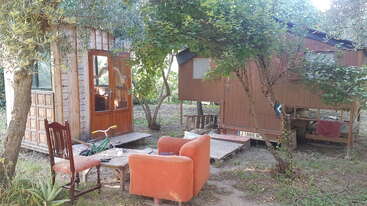 Two small wooden cabins sit among trees. In front, mismatched outdoor furniture, including a red armchair and wooden chairs, creates a cozy, rustic gathering spot.