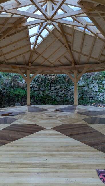Esta imagen muestra un cenador de madera con suelo de madera con motivos geométricos, laterales abiertos y techo transparente, situado frente a un muro de piedra y vegetación.