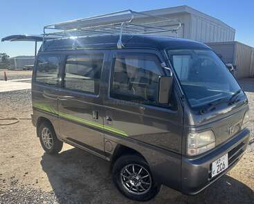 Uma van Honda cinza compacta, com bagageiro no teto e rodas brilhantes, está estacionada ao ar livre perto de prédios industriais em um dia ensolarado com céu limpo.
