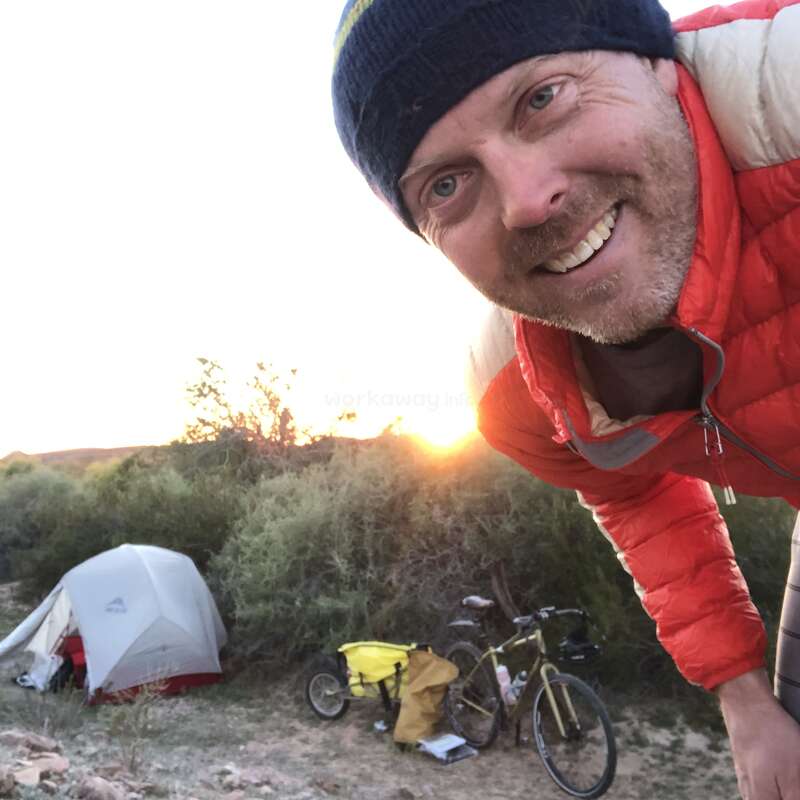 A imagem mostra um homem com uma jaqueta vermelha e um gorro preto, parado em frente a um acampamento com uma barraca, bicicletas e arbustos, em um céu claro.