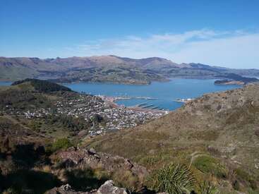 A stunning coastal landscape features green hills, a blue bay with docks and boats, scattered houses, and distant mountains under a clear, bright sky. Beautiful scenery.