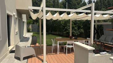 Un patio extérieur moderne comprend une terrasse en bois, une pergola blanche avec un store en tissu, une table de salle à manger avec des chaises, des meubles de salon, des plantes en pot et une pelouse verdoyante.