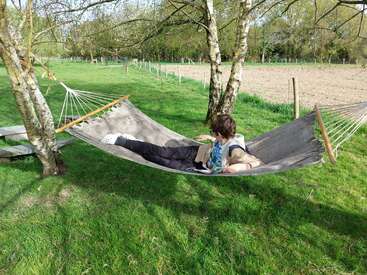 Una persona se relaja en una hamaca colgada entre dos árboles, rodeada de hierba verde, campos y un apacible paisaje rural en un día soleado.