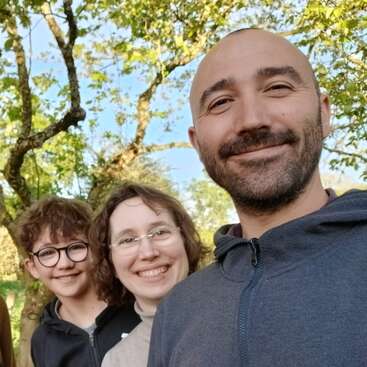 Eine lächelnde Dreiergruppe, darunter ein Kind, steht an einem sonnigen Tag draußen unter grünen Bäumen und verbreitet gemeinsam eine fröhliche, entspannte und heitere Stimmung.