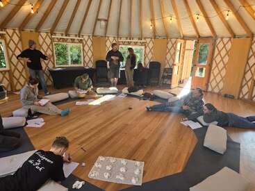 Huit personnes participent à un atelier détendu à l'intérieur d'une yourte confortable, assises sur des coussins, écrivant, conversant et interagissant dans un espace circulaire en bois bien éclairé.