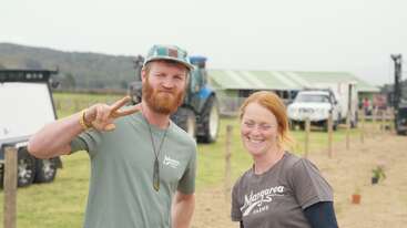 Deux personnes sourient en plein air dans une ferme, portant des chemises "Mangara Farms". L'homme fait un signe de paix. Des tracteurs, des véhicules et des bâtiments agricoles apparaissent à l'arrière-plan.