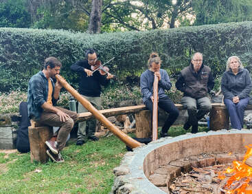 Cinq personnes sont assises autour d'un feu de camp en plein air. Deux jouent du didgeridoos, une joue du violon, tandis que les autres écoutent tranquillement, entourés de verdure et d'arbres.