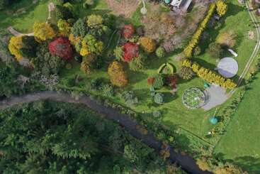 Vue aérienne d'un parc luxuriant avec des arbres d'automne colorés, des chemins sinueux, une petite rivière, des pelouses ouvertes, une structure circulaire et une serre géométrique. Paysage paisible.