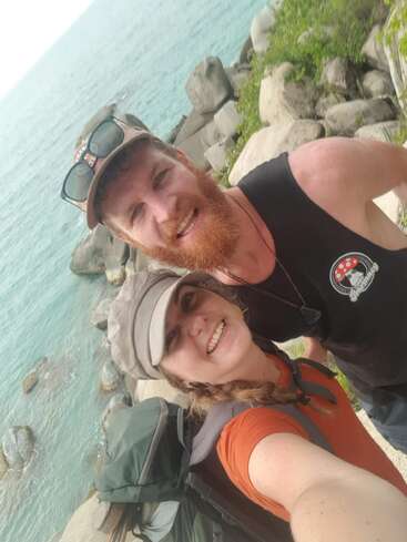 Un couple souriant prend un selfie sur un terrain rocheux au bord de l'océan bleu. Ils portent des vêtements et des chapeaux décontractés, et profitent manifestement d'une aventure pittoresque ensemble.