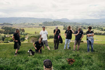 Un groupe de personnes se tient debout et discute sur une colline herbeuse avec des terres agricoles et des montagnes en arrière-plan. Une personne est agenouillée à côté d'un chien noir et blanc.