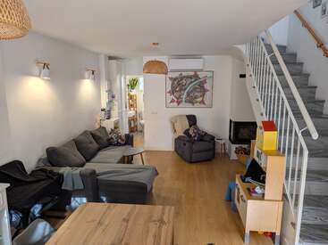 Ein gemütliches, modernes Wohnzimmer mit einem grauen Ecksofa, einem Sessel, Holzboden, einer Treppe, Kinderspielzeug, Wanddekoration, Hängeleuchten und einem kleinen Esstisch.