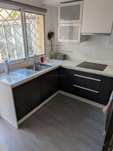 Diese moderne Küche verfügt über große Fenster mit Sicherheitsgittern, schlichte dunkle Schränke, weiße Arbeitsplatten, eine Doppelspüle, ein Elektrokochfeld und eine minimalistische Einrichtung, zu der auch eine kleine Pflanze gehört.