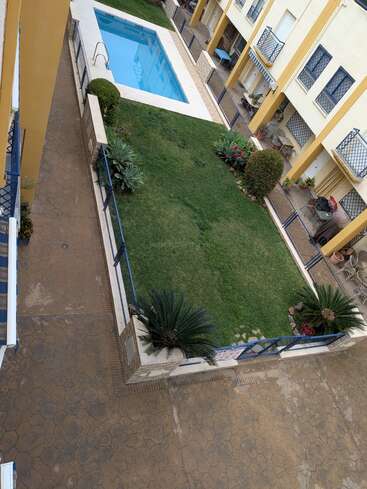 Dieses Bild zeigt den Innenhof einer Wohnanlage mit einem kleinen Swimmingpool, einem grünen Rasen, Zierpflanzen, Sträuchern, Gartenmöbeln und gelb gestrichenen Gebäuden mit blauen Geländern.
