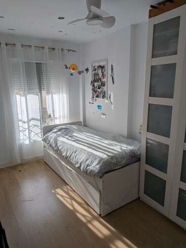 Ein gemütliches, sonnendurchflutetes Schlafzimmer mit einem Einzelbett, weißen Wänden, Holzboden, Kleiderschrank, Deckenventilator, großem Fenster, transparenten Vorhängen, einem Planeten-Mobile und einer Fotocollage.