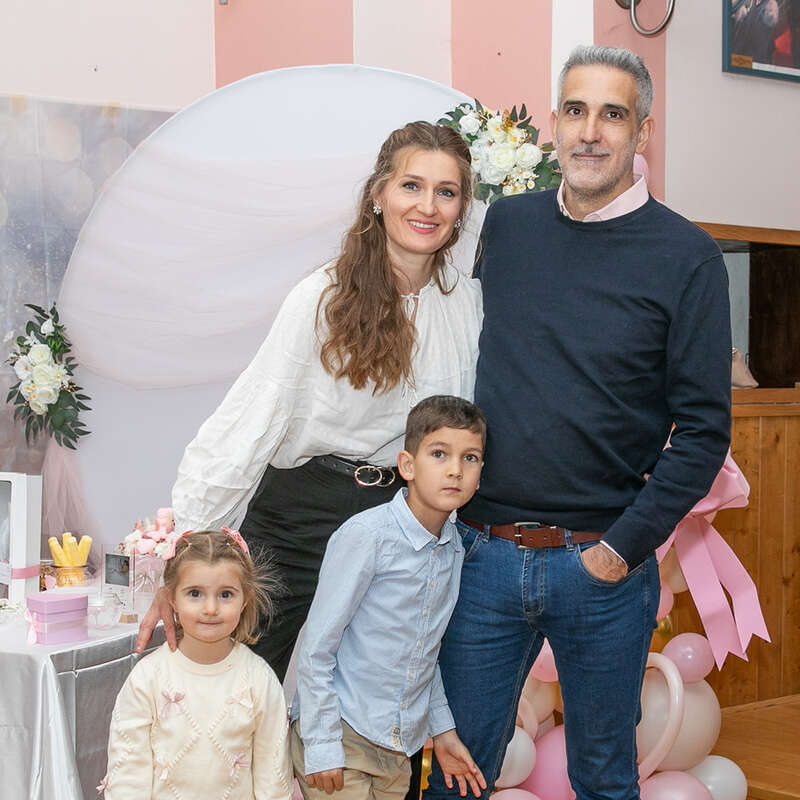 Eine lächelnde vierköpfige Familie posiert drinnen bei einer Veranstaltung, die mit rosa und weißen Luftballons geschmückt ist. Die Eltern stehen hinter ihrem kleinen Sohn und ihrer kleinen Tochter.