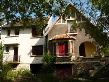 The image depicts a two-story house with a brown roof, white walls, and red doors, surrounded by trees and bushes on a sunny day.