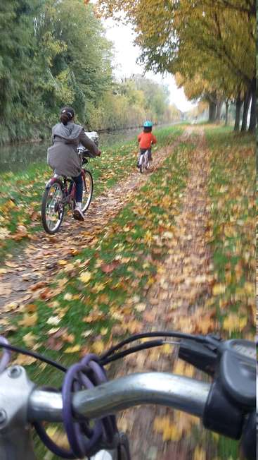Das Bild zeigt zwei Kinder auf Fahrrädern auf einem mit Laub bedeckten Weg neben einem Fluss, mit Bäumen, die den Weg säumen, und einer ruhigen Atmosphäre.