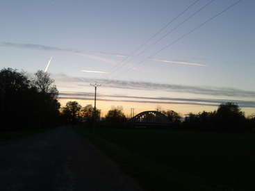 Das Bild zeigt eine ruhige ländliche Landschaft bei Sonnenuntergang mit einer Brücke, einem Telefonmast, Bäumen und Stromleitungen vor einem strahlenden Himmel mit Wolken.