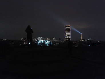 Das Bild zeigt eine nächtliche Stadtlandschaft mit einem hohen Gebäude, von dessen Spitze ein Lichtstrahl ausgeht, vor dem Hintergrund eines dunklen Himmels.