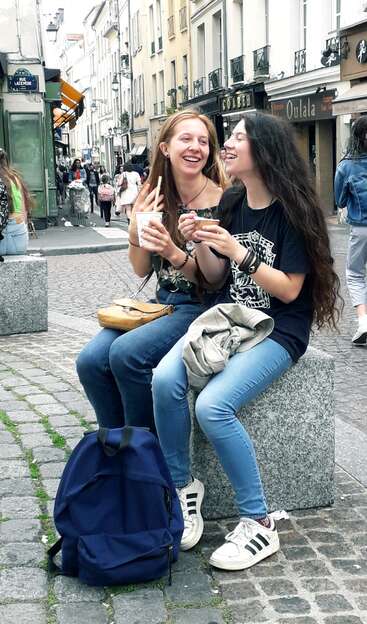 Zwei junge Frauen sitzen lachend und mit Getränken in der Hand auf einer Steinbank. Im Hintergrund sind eine Straße und Gebäude zu sehen, die eine sorglose Atmosphäre ausstrahlen.