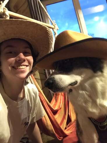 Eine lächelnde Person und ein Hund tragen beide Hüte in einem Haus. Sie scheinen glücklich zu sein und sitzen zusammen vor einem Fenster mit einer Hängematte hinter ihnen.