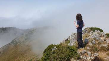 Eine Person mit langen Haaren steht auf einem felsigen Berggipfel, umgeben von Nebel und Wolken, und blickt in die Ferne. Die Vegetation und das zerklüftete Gelände sind sichtbar.
