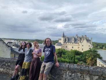 Vier Menschen stehen lächelnd vor einer Steinmauer, mit einer historischen Burg, üppigem Grün und einem Fluss im malerischen Hintergrund unter bewölktem Himmel.