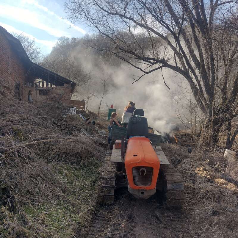 The image depicts a tractor with two people on board, navigating through a muddy path amidst a pile of debris and smoke in the background.