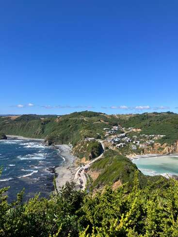 Eine atemberaubende Küstenlandschaft mit sanften grünen Hügeln, schroffen Klippen, Wellen, die an die Küste schlagen, einer kurvenreichen Straße, vereinzelten Häusern und einem strahlend blauen Himmel darüber.