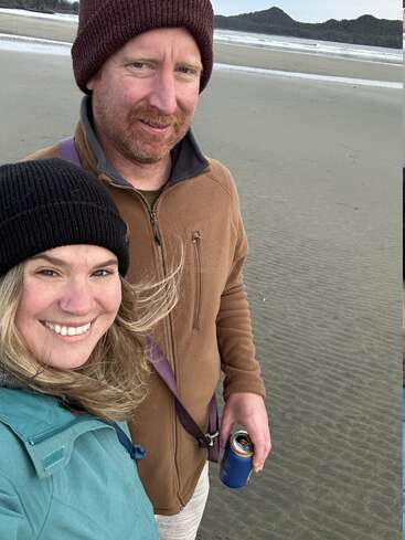 Ein lächelndes Paar mit Wintermützen und Jacken steht an einem Sandstrand. Der Mann hat eine Dose in der Hand, während hinter ihnen das Meer und entfernte Hügel zu sehen sind.