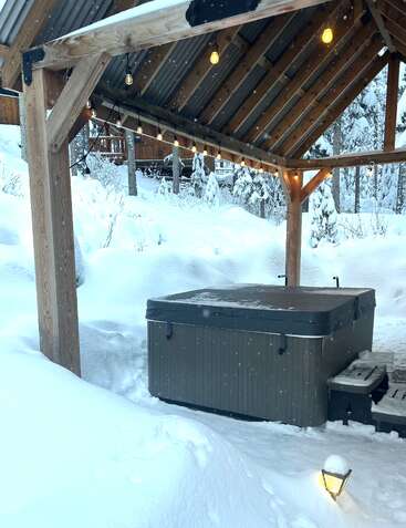 Ein überdachter Whirlpool liegt unter tiefem Schnee unter einem hölzernen Pavillon. Warme Lichterketten leuchten darüber und schaffen einen gemütlichen Rückzugsort im Winter, umgeben von verschneiten Bäumen.