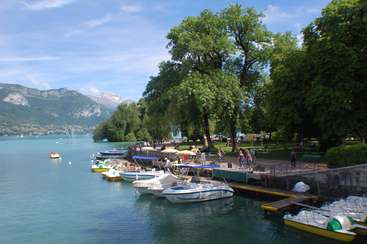 L'image représente une scène de lac sereine avec des bateaux amarrés le long de la rive, entourés d'arbres luxuriants et de montagnes en arrière-plan, sous un ciel bleu avec des nuages blancs.