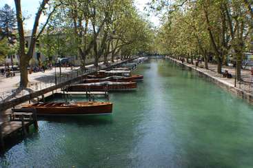 L'image représente une scène sereine de canal, avec des bateaux, des arbres et une promenade, avec une rangée de bâtiments bordant le côté opposé de la voie d'eau.