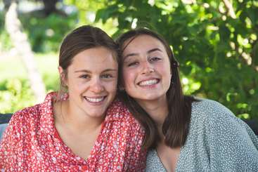 L'image montre deux femmes posant pour une photo, la femme de gauche portant un haut blanc et la femme de droite portant un haut noir, toutes deux les bras croisés.