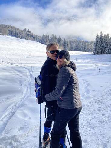 L'image représente un couple enlacé dans un paysage enneigé, l'homme portant des lunettes de soleil et tenant des bâtons de ski, et la femme portant un bonnet et des lunettes de soleil.