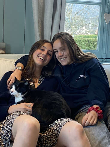 L'image représente deux femmes assises sur un canapé, l'une tenant un chat noir et blanc, avec une fenêtre et un jardin visibles à l'arrière-plan.