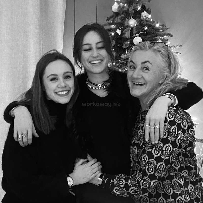 L'image est une photographie en noir et blanc de trois femmes posant devant un arbre de Noël, la femme de gauche portant un pull sombre et les deux autres des hauts sombres.
