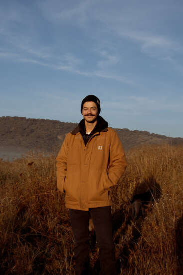 A man wearing a brown jacket and black beanie stands smiling in a grassy field with mountains in the background during a clear, sunny day. Dog partially visible.