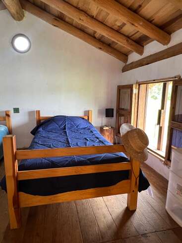 A cozy rustic bedroom with wooden beams, a single bed with dark blue bedding, a straw hat on the bedpost, open window, and natural light.