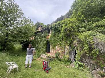 Ein rustikales Steinhaus, das mit dichtem grünem Efeu bewachsen ist, steht in einem üppigen, verwilderten Garten. Man sieht zwei Menschen, einen weißen Stuhl und einen Rucksack.
