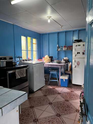 Esta é uma cozinha simples, com paredes azuis, piso de azulejo, fogão, geladeira, máquina de lavar, uma mesinha, um banquinho, um refrigerador portátil, prateleiras abertas e iluminação fluorescente.
