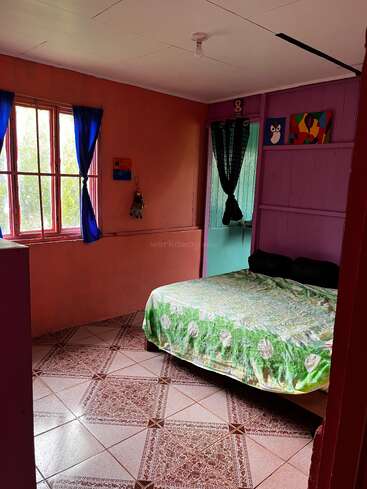 Um quarto aconchegante com paredes vermelhas e roxas, piso de azulejo, colcha verde, duas janelas com cortinas azuis e pretas e decorações coloridas nas paredes.