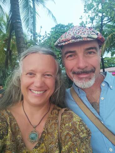 Un couple souriant prend un selfie en plein air. La femme porte un grand collier à pendentifs et l'homme une casquette à motifs. Des palmiers et de la verdure les entourent.