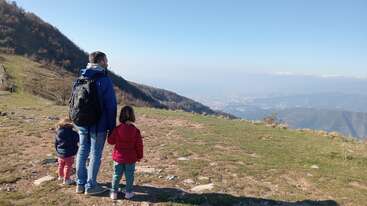 A man and two young children stand on a grassy hilltop, gazing at a distant city below, surrounded by mountains under a clear, blue sky.