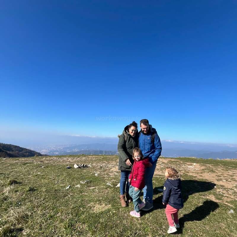 A family of four stands outdoors on a grassy hill, enjoying a sunny day with a clear blue sky and mountains visible in the far distance.