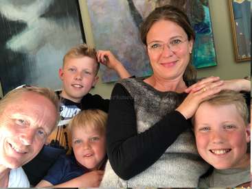 A imagem mostra uma família de cinco pessoas posando para uma selfie, com a mãe e o pai à esquerda e três filhos à direita, todos sorrindo e olhando para a câmera.
