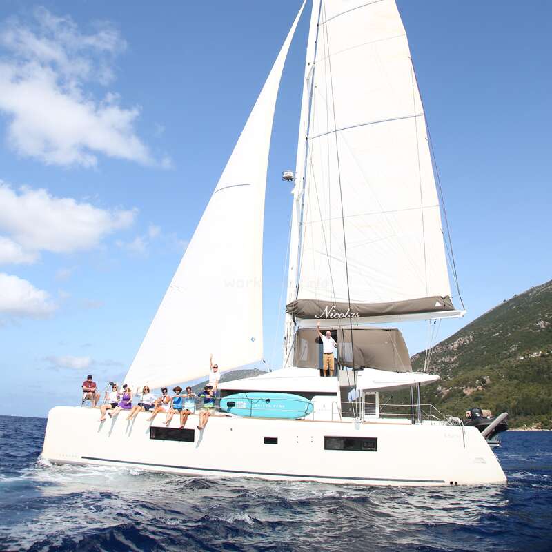L'image représente un grand catamaran blanc avec un groupe de personnes à bord, naviguant sur l'océan par une journée ensoleillée, avec un littoral montagneux en arrière-plan.