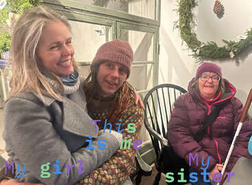 L'image montre trois femmes assises ensemble, souriantes, avec une couronne de Noël sur le mur derrière elles. Elle a probablement été prise pendant la période des fêtes de fin d'année.